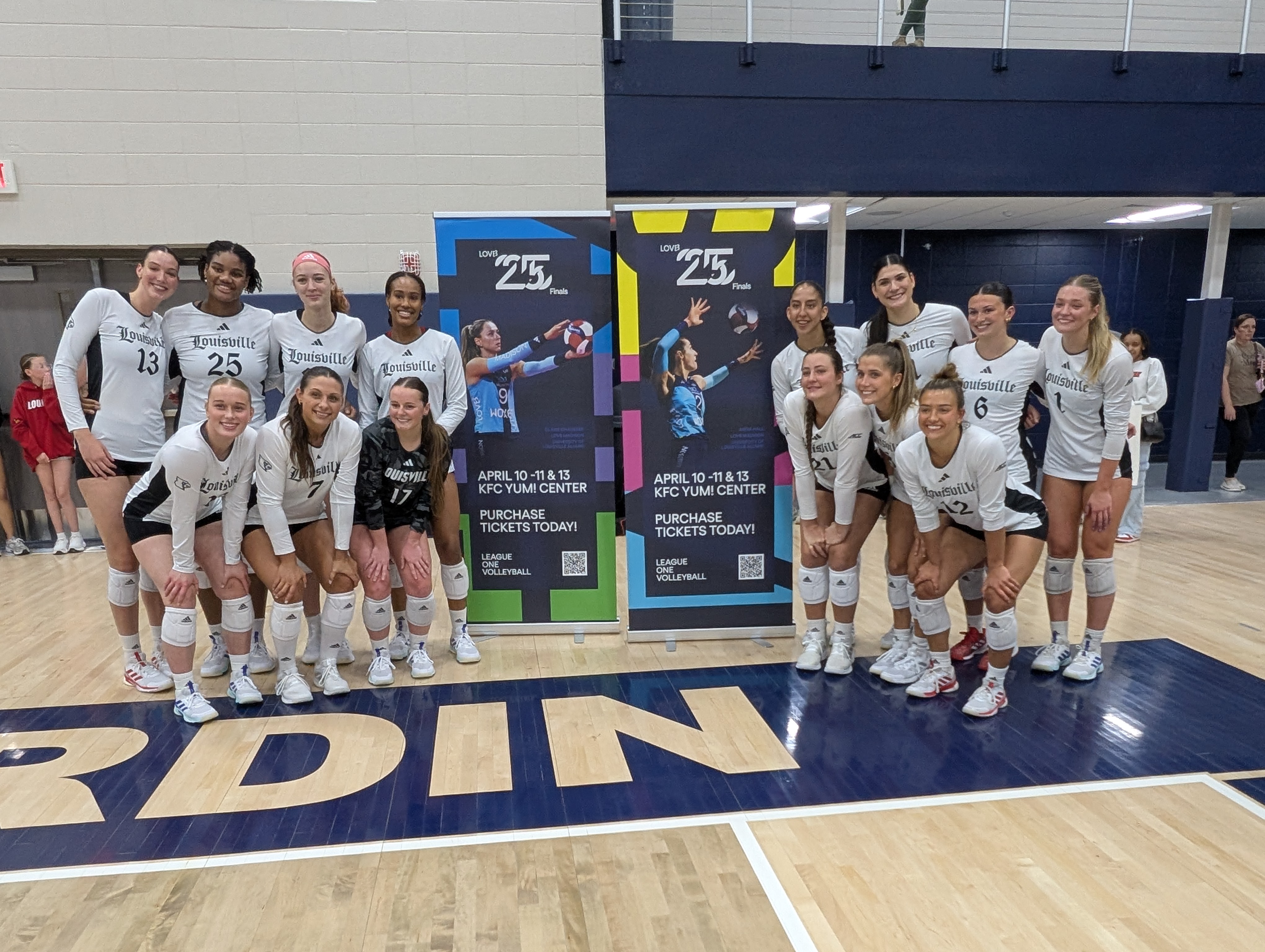 The UofL team gathers around banners featuring alumnae Claire Chaussee and Anna "Stevie" Hall (nee Stevenson) advertising the LOVB finals event at the KFC Yum! Center, April 10-11 & 13th